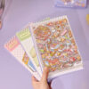 Carnet à spirales contenant 20 scènes de stickers isométrique 3D à composer soi-même sur le thème des boutiques kawaii et mignonnes. Activité créative pour adulte pour gérer le stress.